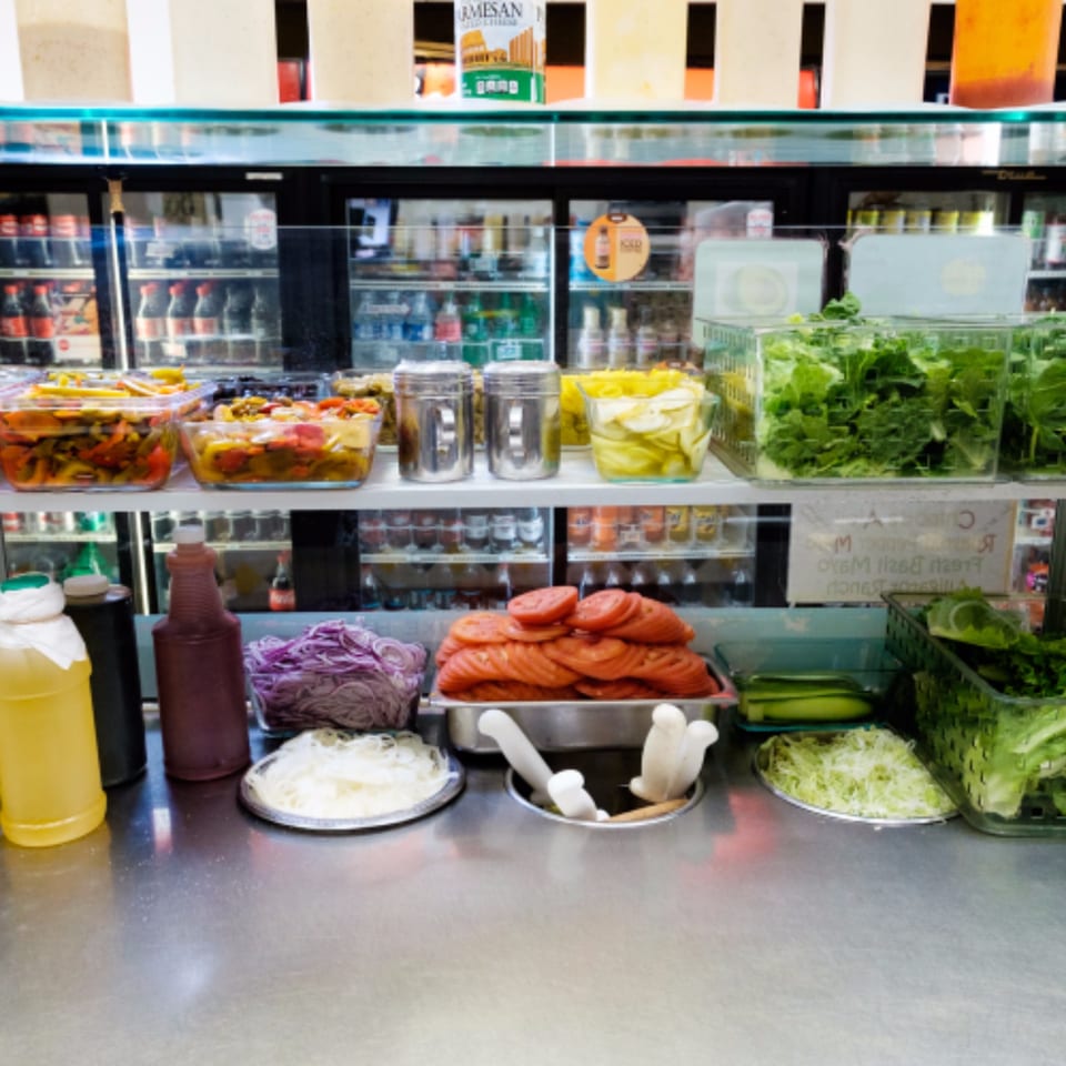 A sandwich bar with various fresh toppings like tomatoes, lettuce, onions, and pickled vegetables.